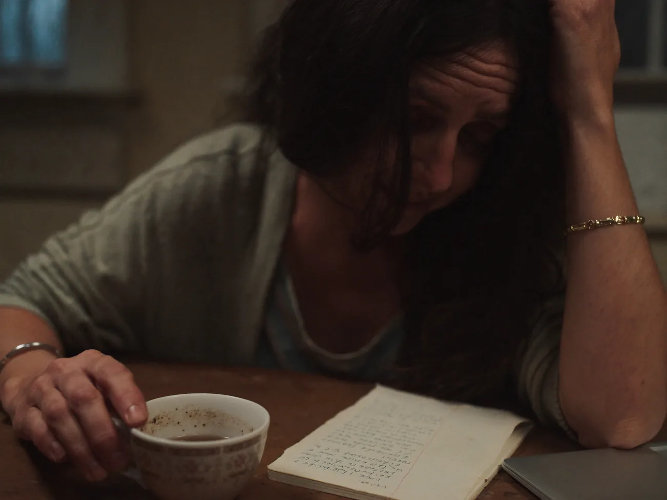 Dunkelhaarige Frau am Küchentisch am Abend, eine Hand im Haar, Blick gesenkt auf ein Notizbuch — müde, alltagsnah. Daneben eine Tasse Tee und ein Laptop.