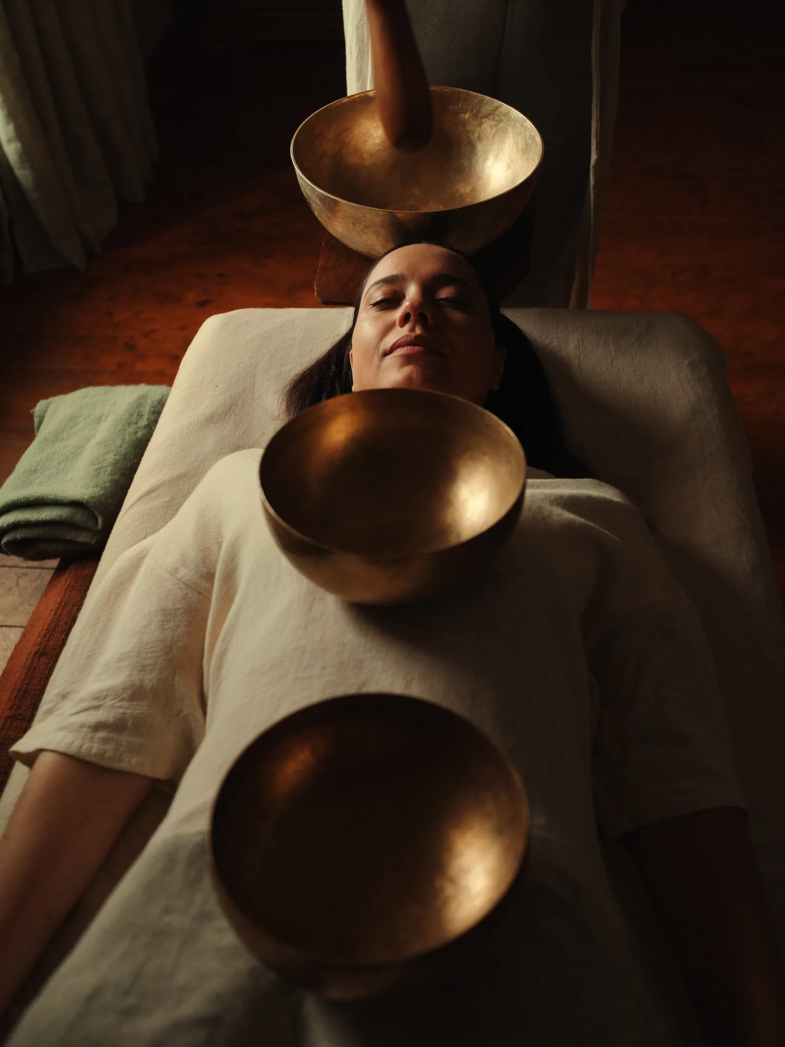 Klangmassage-Sitzung: Frau liegt entspannt auf einer hellbeigen Massageliege, Augen geschlossen, drei goldene Messing-Klangschalen — eine auf dem Brustbein, eine auf dem Unterbauch, eine hinter dem Kopf. Eine Hand spielt eine Schale mit Holzschläger an.