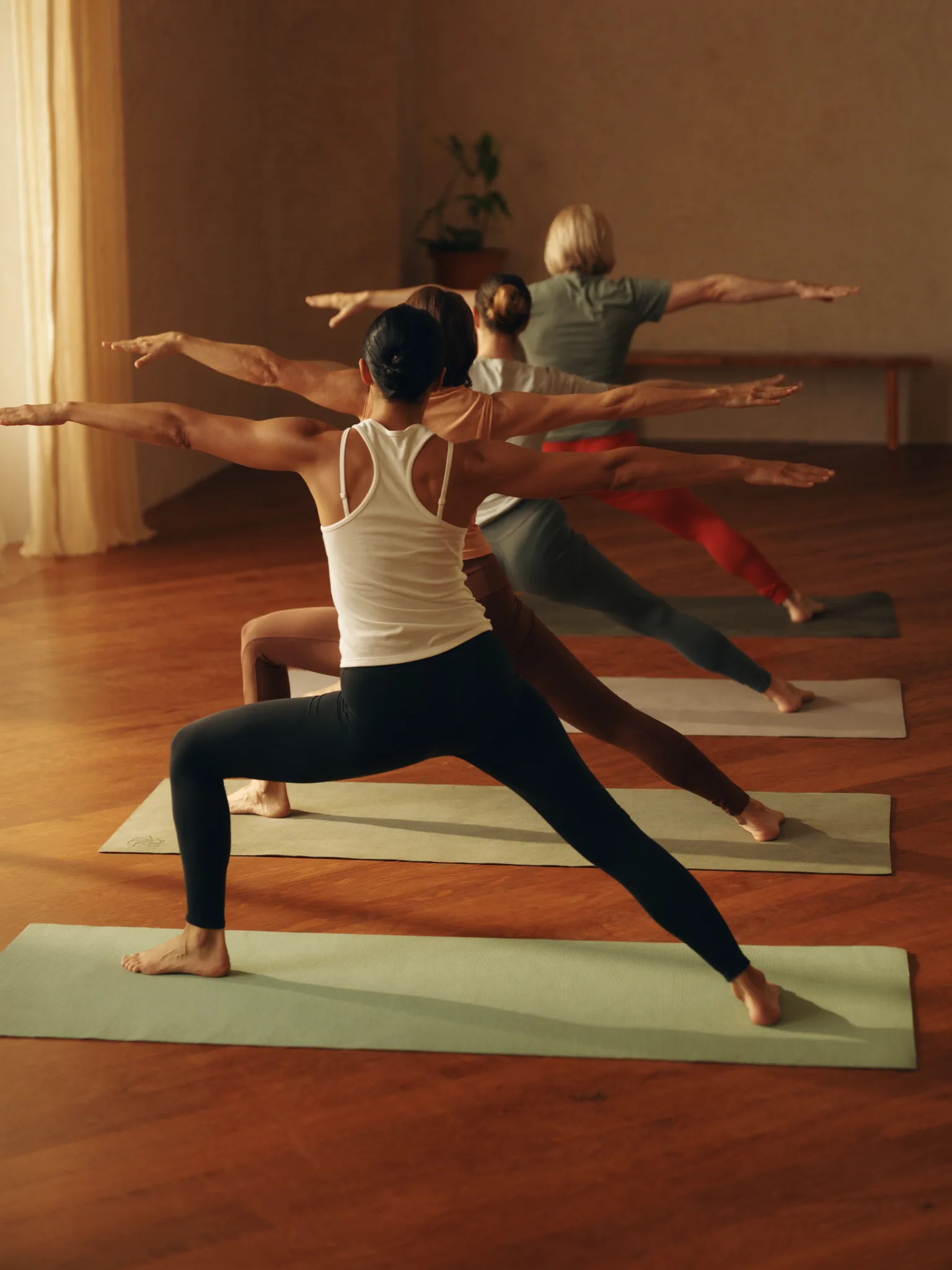Vinyasa-Yogastunde im warmen Studio: vier Praktizierende von hinten in der Pose Krieger 2, sage-farbene Yogamatten, Holzboden, weiches Tageslicht von links.